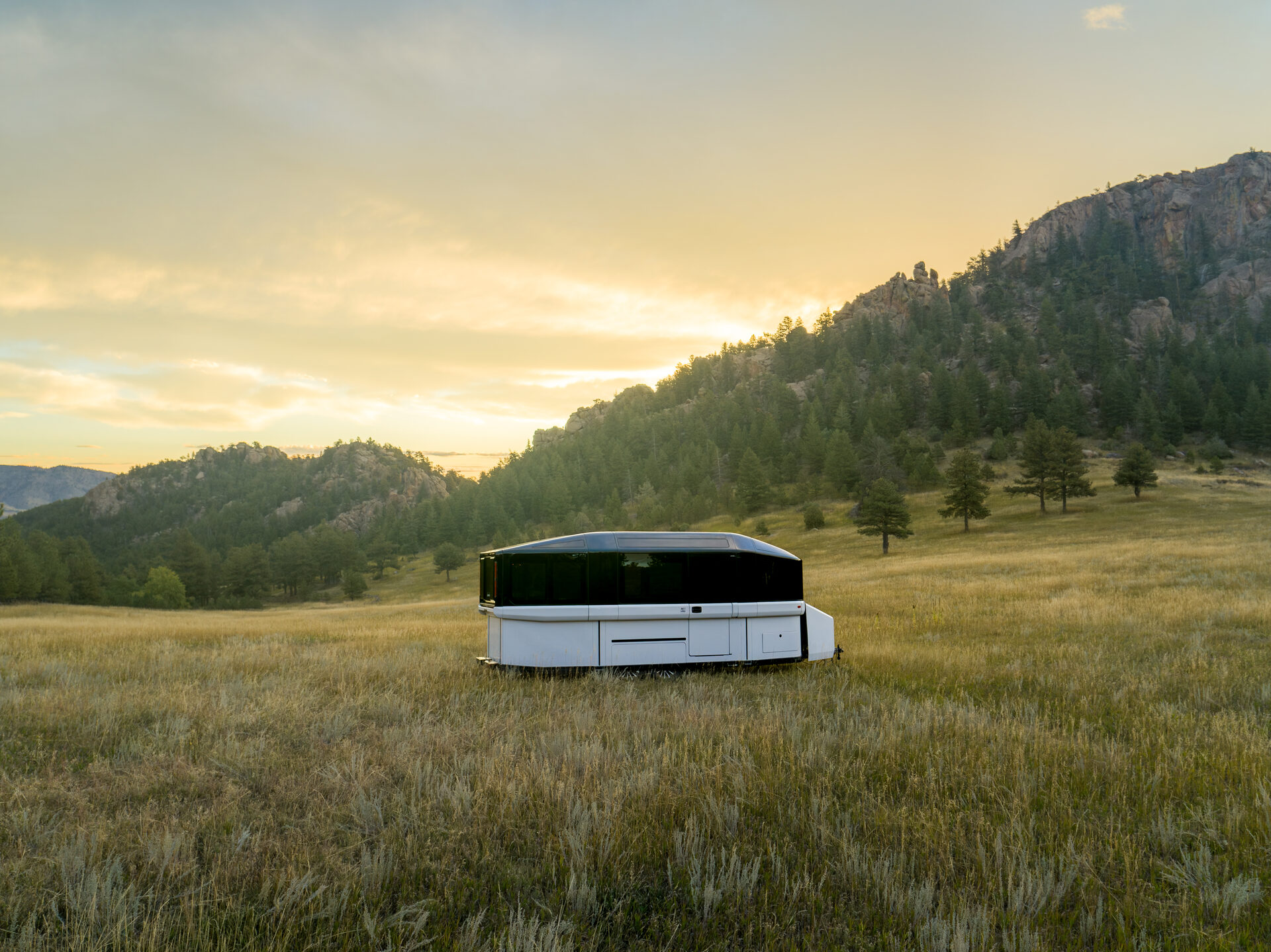 The Lightship AE1 in road mode, parked in an open mountain meadow at sunset with pine-covered rocky hills behind it
