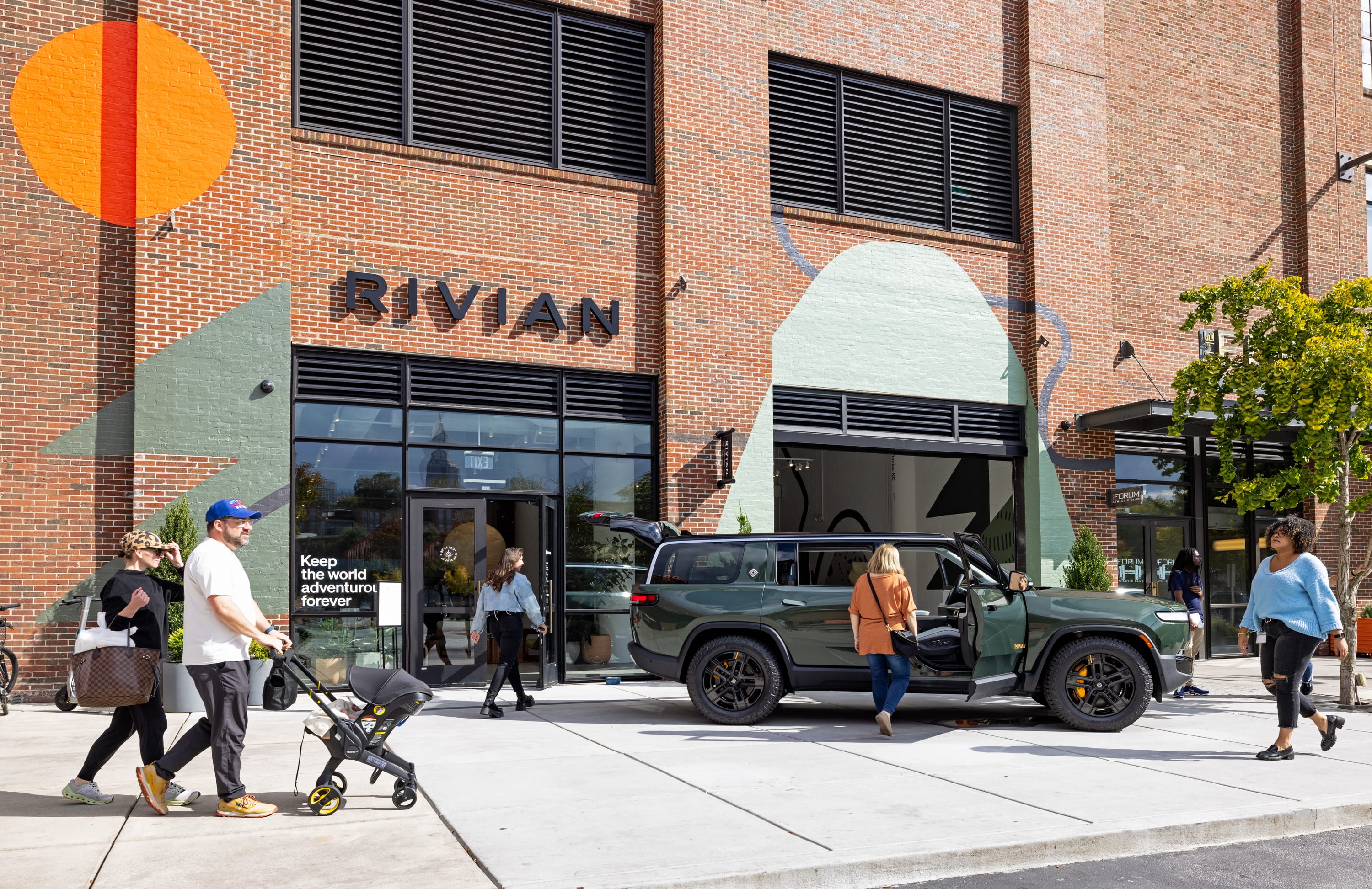 Pedestrians walking past a Rivian retail location in Atlanta, with a green R1S parked outside and the tagline Keep the world adventurous forever visible in the window